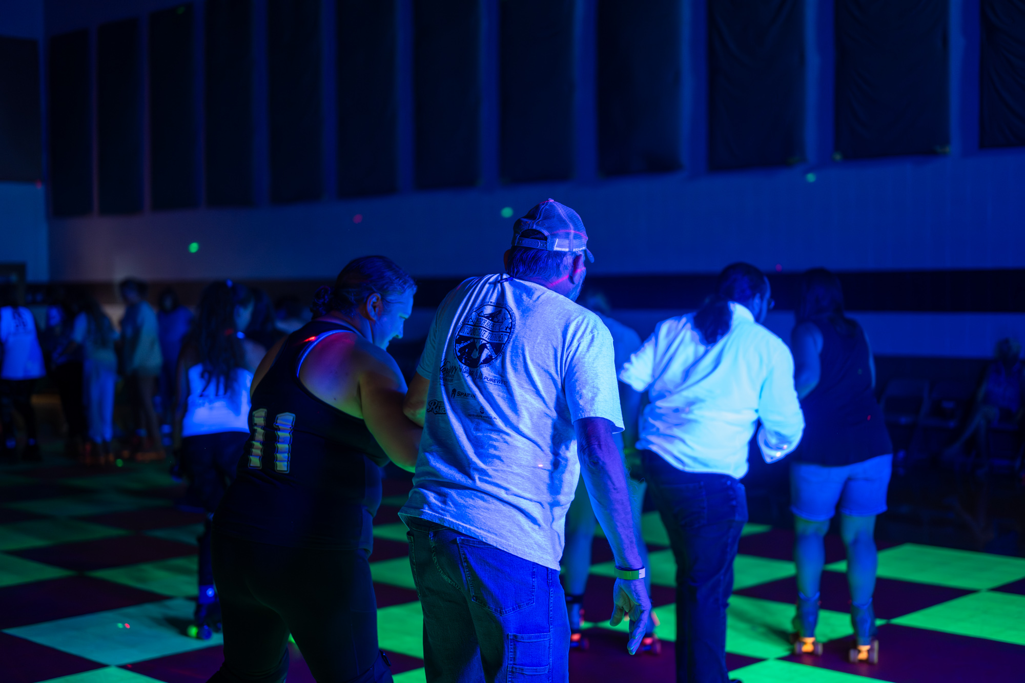 Dark room with green and blue neon lights and people roller skating on a checkerboard floor