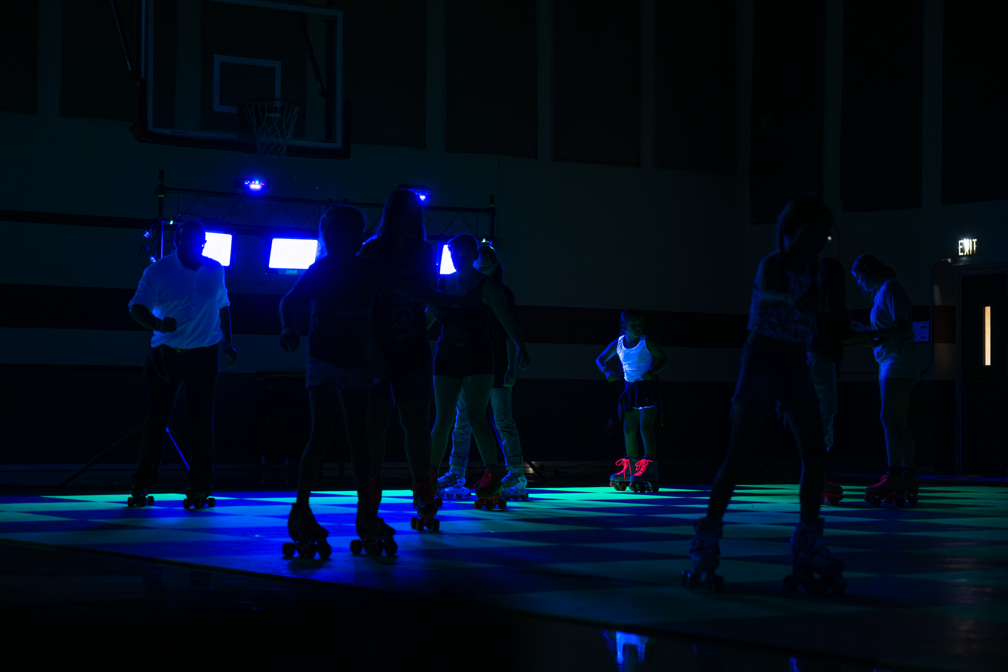 Dark room with green and blue neon lights and people roller skating on a checkerboard floor