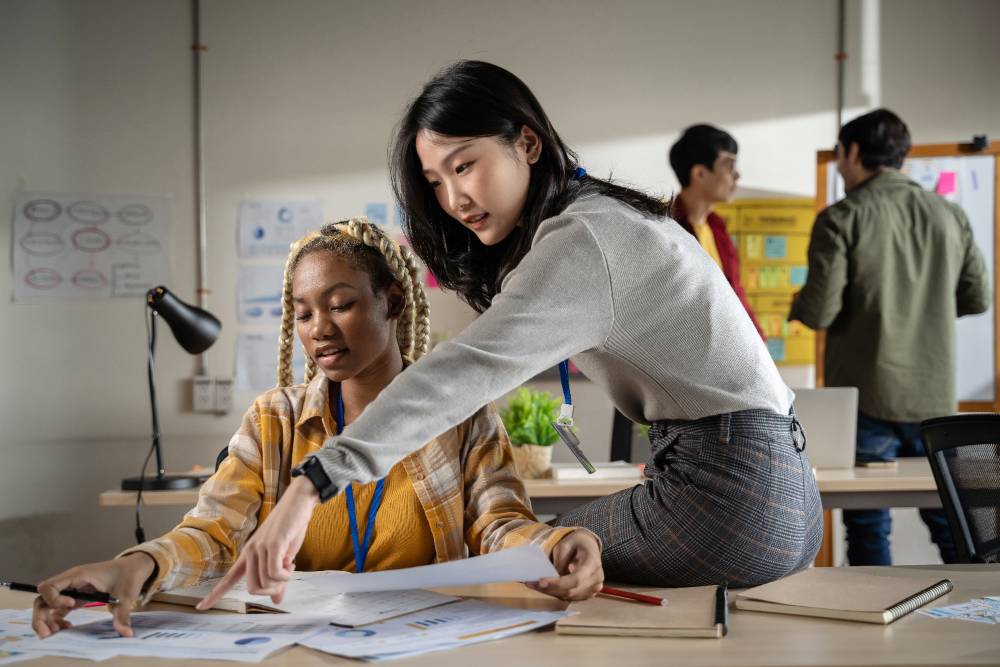 A female student assisting another as two male students discuss something in the background