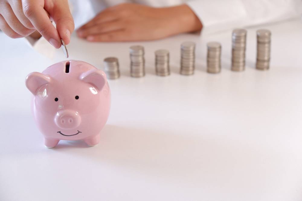 Person Inserting Coins In Piggy Bank With Stack Of Coins Over The Desk
