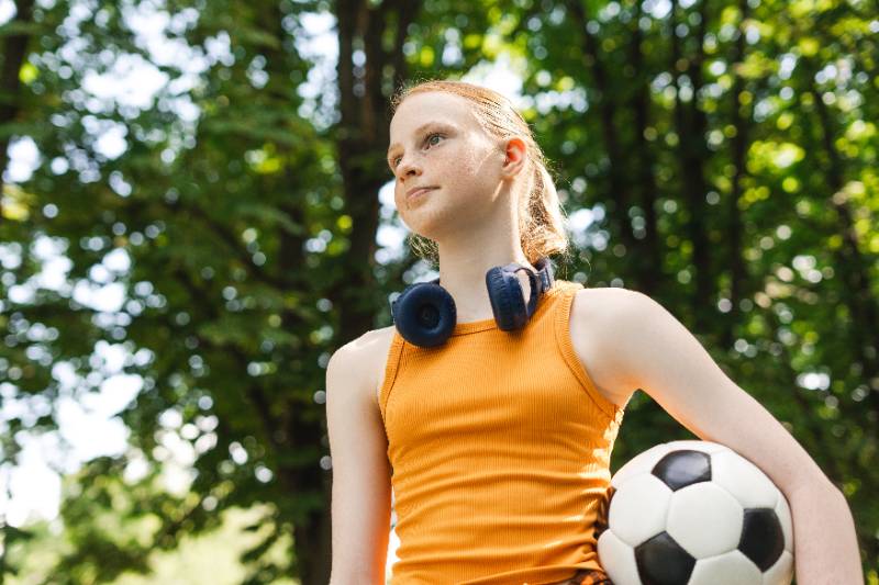 Red hair teenager girl posing with headphones holding a soccer ball at the park